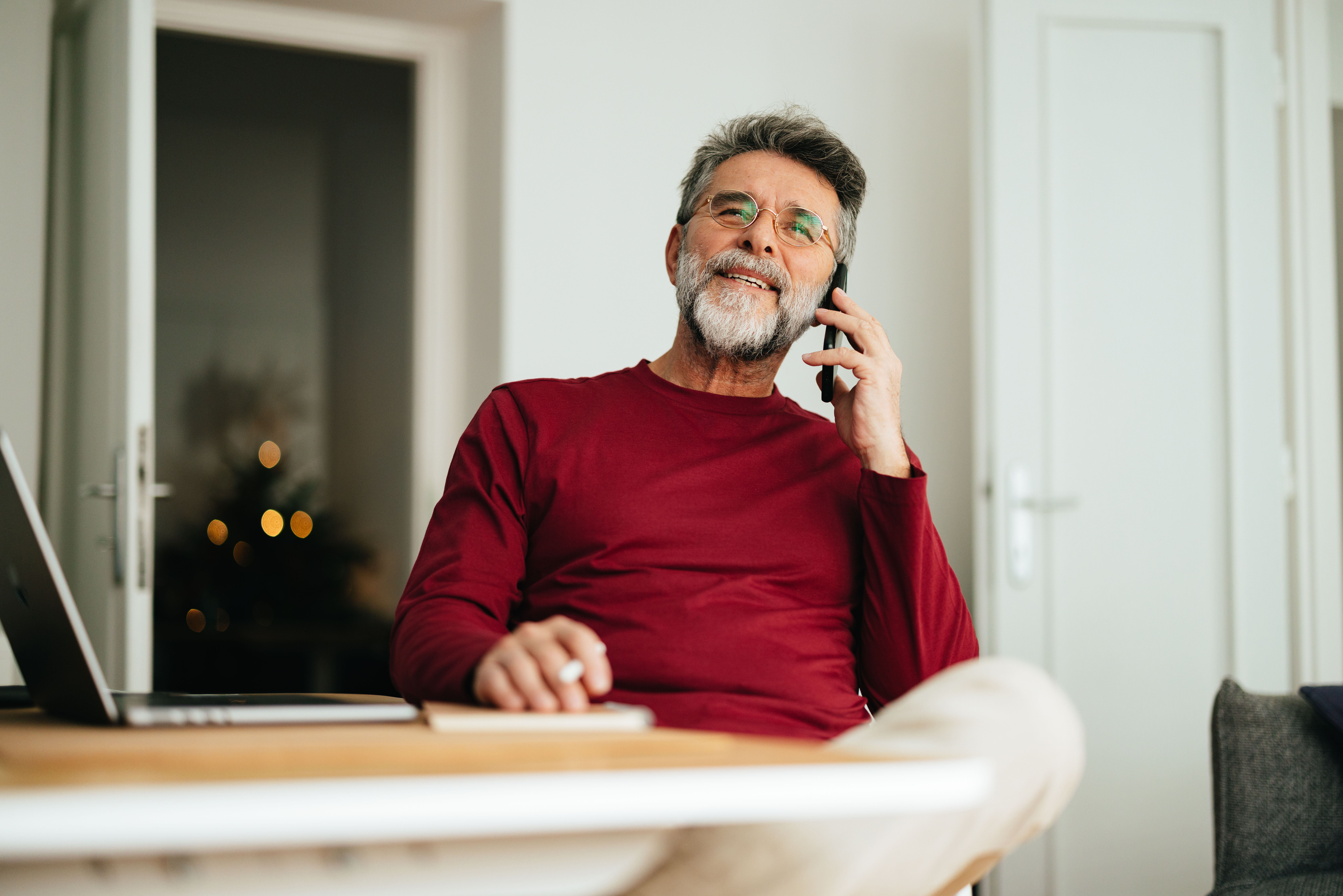 Low angle view of smiling mature business man speaking on the phone while sitting at home office desk with laptop computer, notebook and pen.